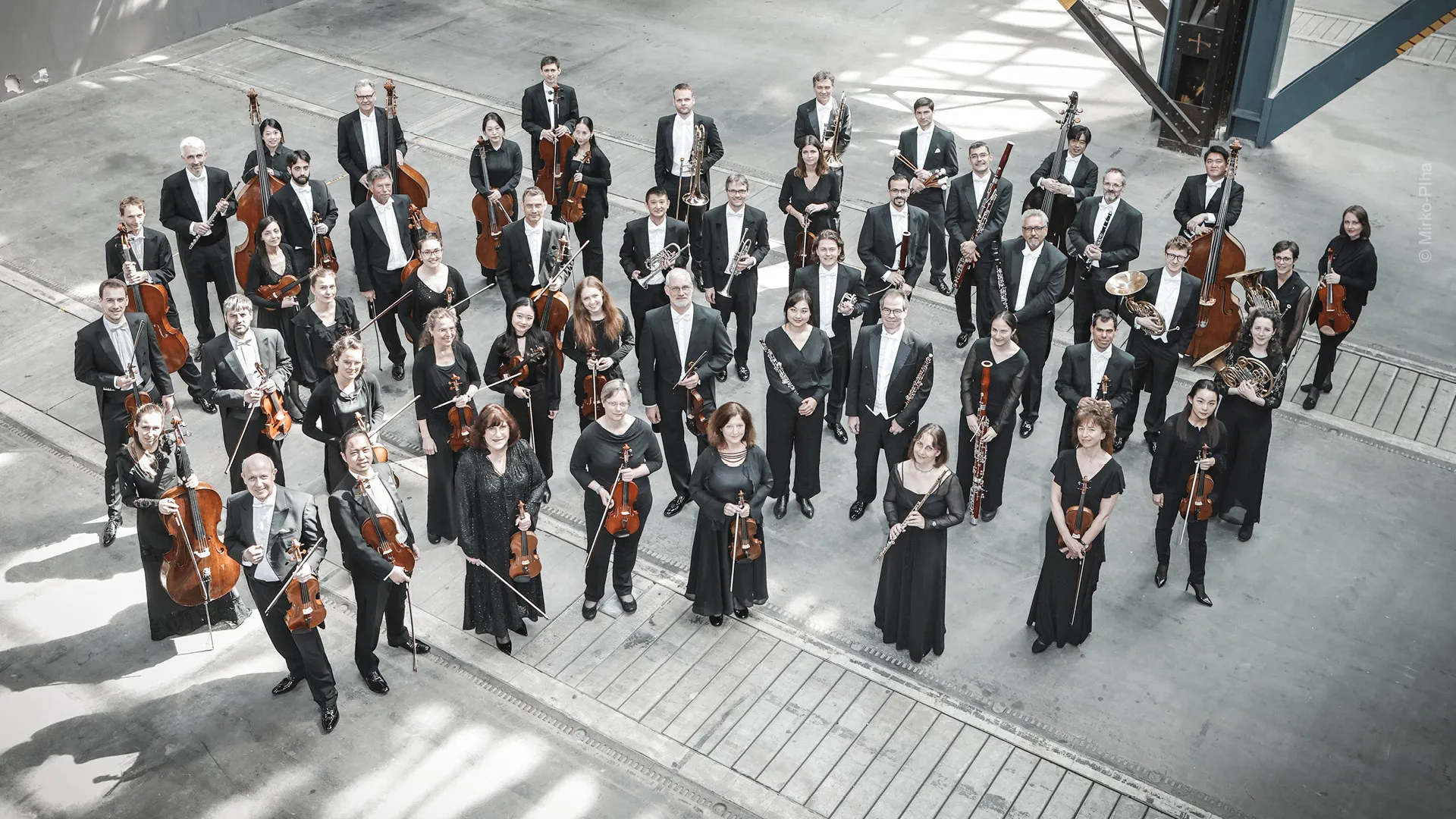 Großes Orchester in Schwarz steht in heller Halle, Musiker mit Streich- und Blasinstrumenten blicken zur Kamera.
